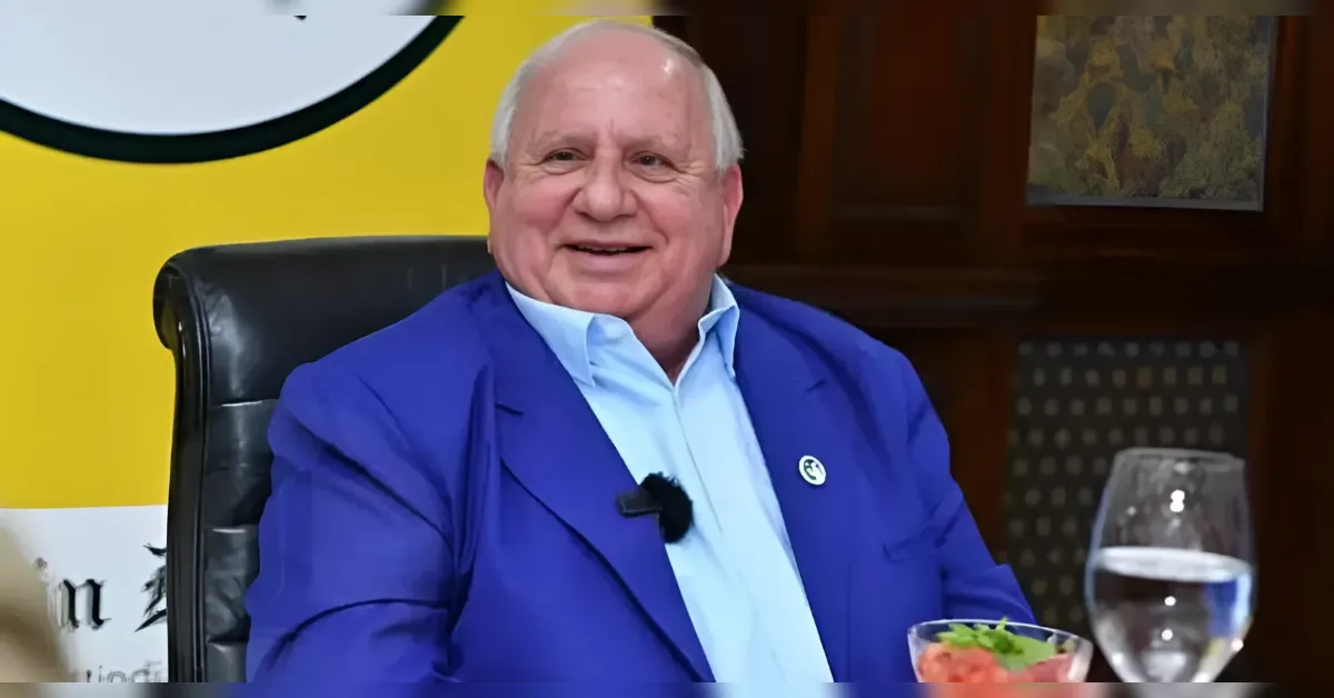 Smiling older man in a blue blazer and light blue shirt sits at a table with a microphone, a glass of water, and a salad in front of him.
