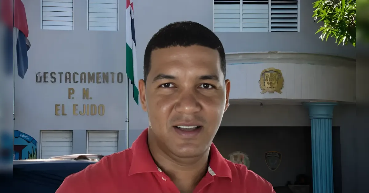 Man in a red polo shirt speaking outdoors in front of a police detachment building labeled 'DESTACAMENTO P.N. EL EJIDO' with flags nearby.