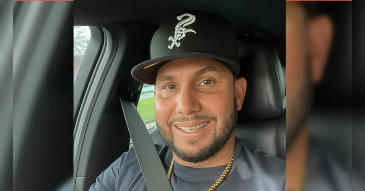 Smiling man wearing a black cap with a logo, seated in a car and buckled up in a seatbelt.