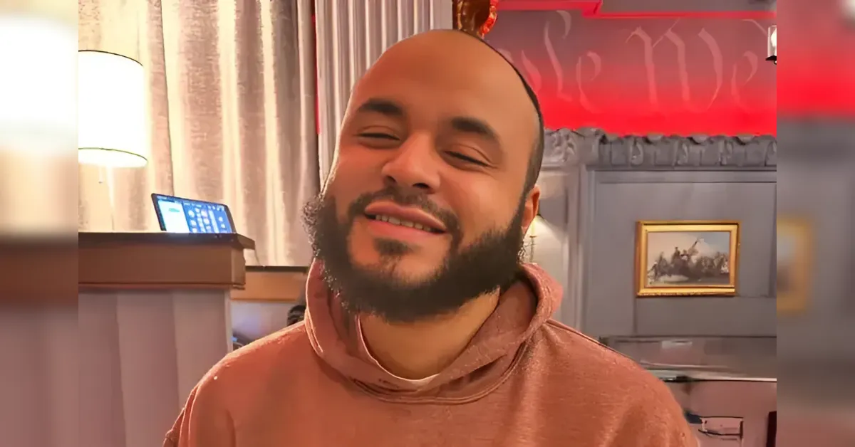 Man with a beard wearing a pink hoodie smiles with eyes closed in a cozy room.