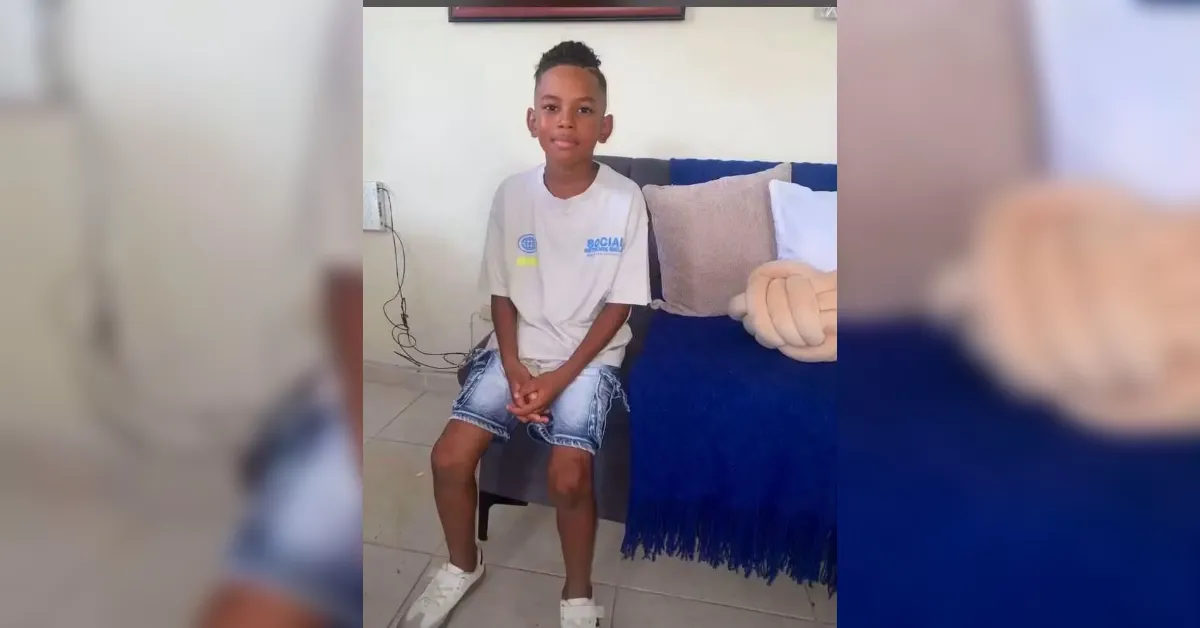 Young boy sits on a small stool in a living room, wearing a gray T‑shirt, denim shorts, and white sneakers, with a blue blanket on a sofa behind him.