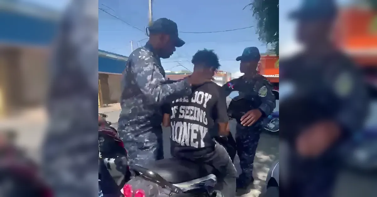 Man on a motorcycle being approached by two uniformed officers on a sunny street; one officer grips his arm while the other stands nearby, a third officer in the background.