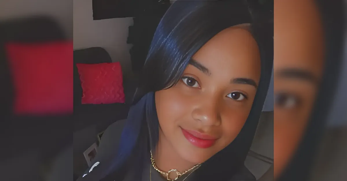 Young woman wearing a dark hijab smiles warmly at the camera indoors, with a pink cushion in the background.