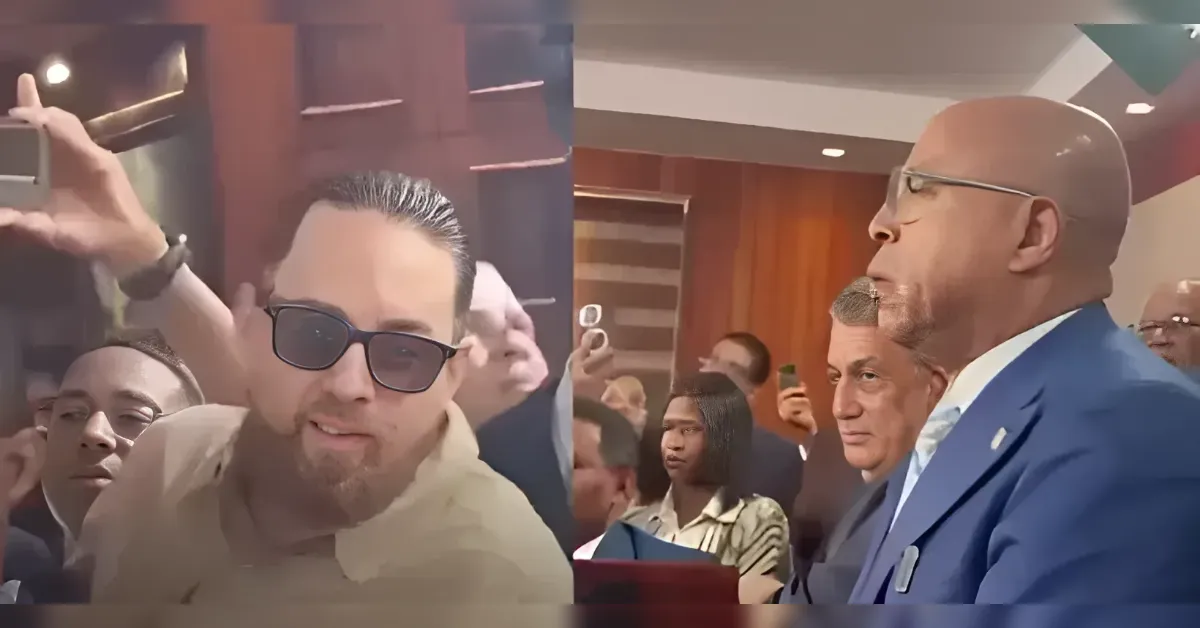 Person wearing sunglasses takes a selfie amid a crowded formal event; a man in a blue suit speaks to the crowd on the right.