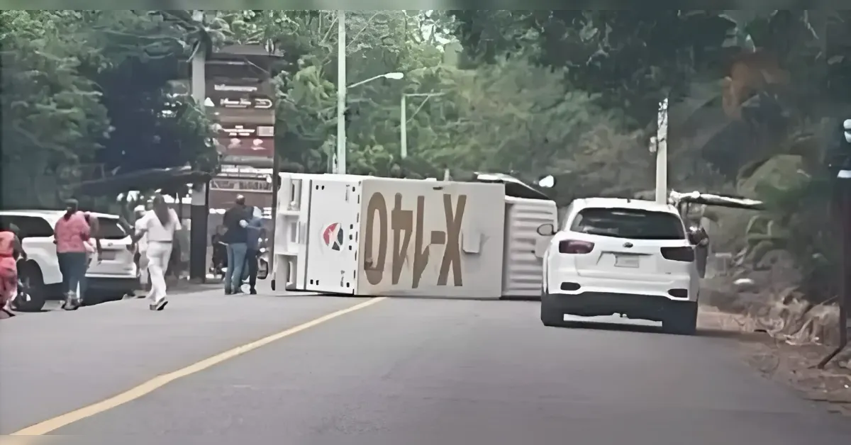Overturned white box truck blocks a two-lane road as bystanders and parked cars watch the scene.