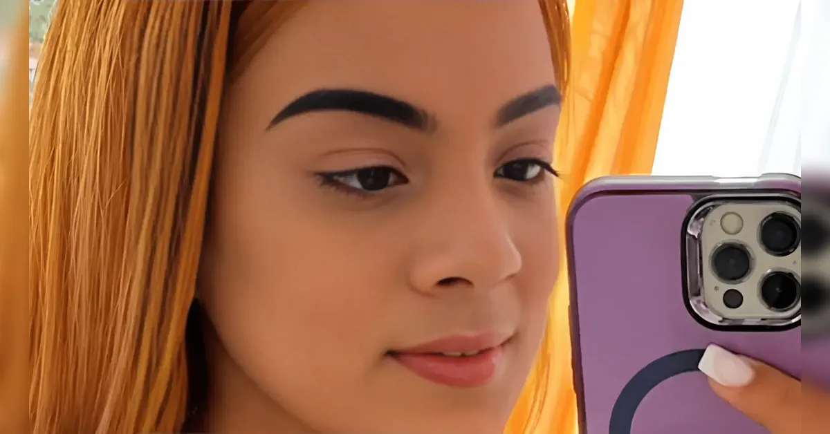Close-up selfie of a person with orange hair holding a purple smartphone in front of a window with orange curtains.