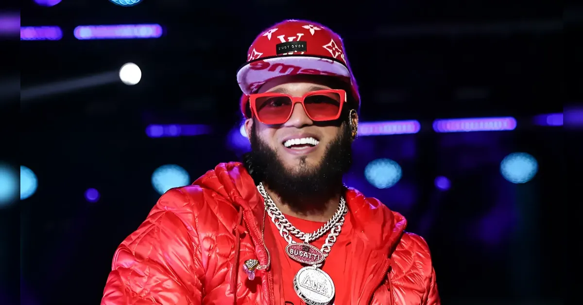 Smiling man with a beard in a red cap, red sunglasses, red puffer jacket, and chunky necklaces in a nightclub setting with purple lighting.