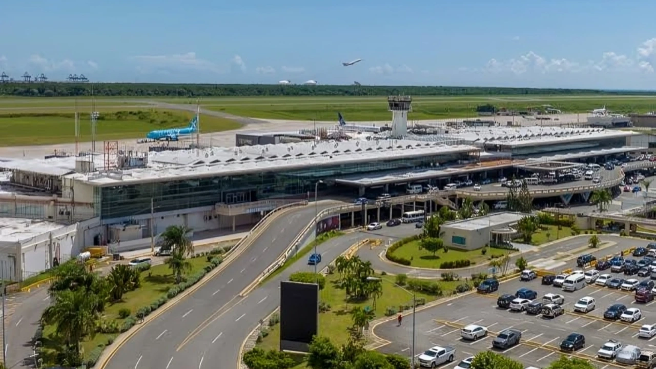 A large airport terminal with a curved road loop in front, numerous parked cars, and green landscape beyond the runway under a blue sky with a plane flying overhead.
