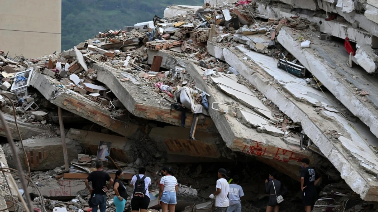 Group of people surveying a collapsed building with large concrete slabs and scattered bricks and debris in an aftermath scene.