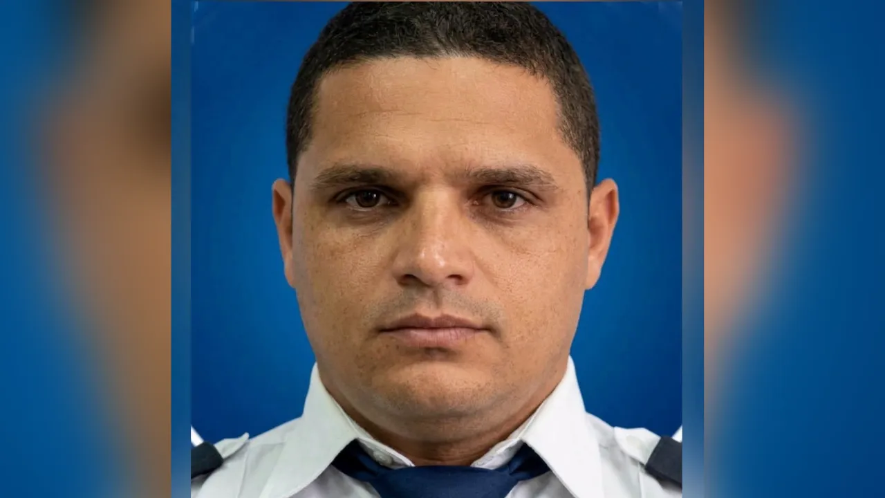 Portrait of a man in a light-colored uniform with a dark blue tie against a blue backdrop, looking straight at the camera with a neutral expression.