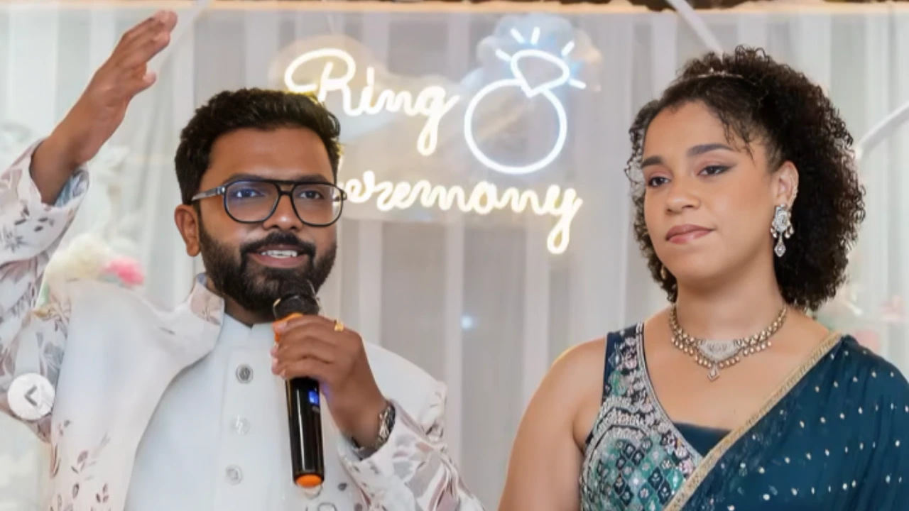 Man with glasses speaks into a microphone beside a woman in a teal sari at a ring ceremony in a festive setting.