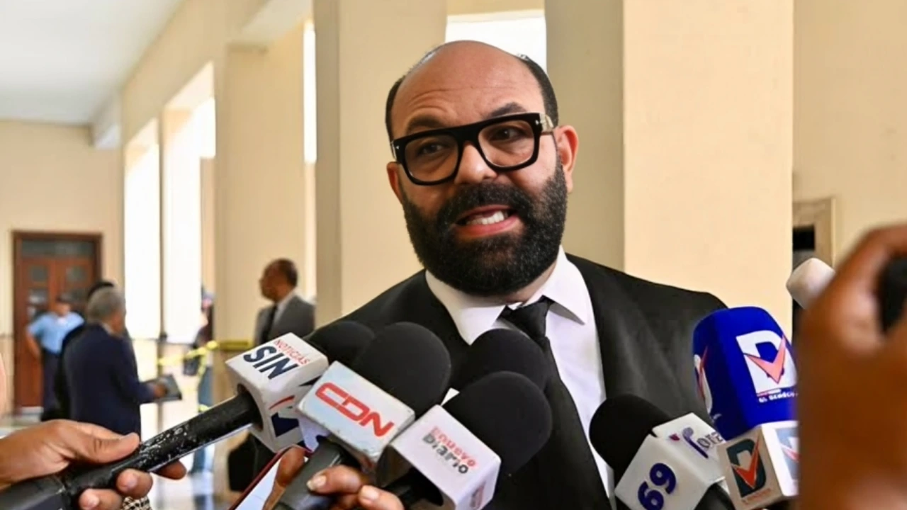 Man in a suit and glasses speaks to reporters, surrounded by many microphones in a hallway.