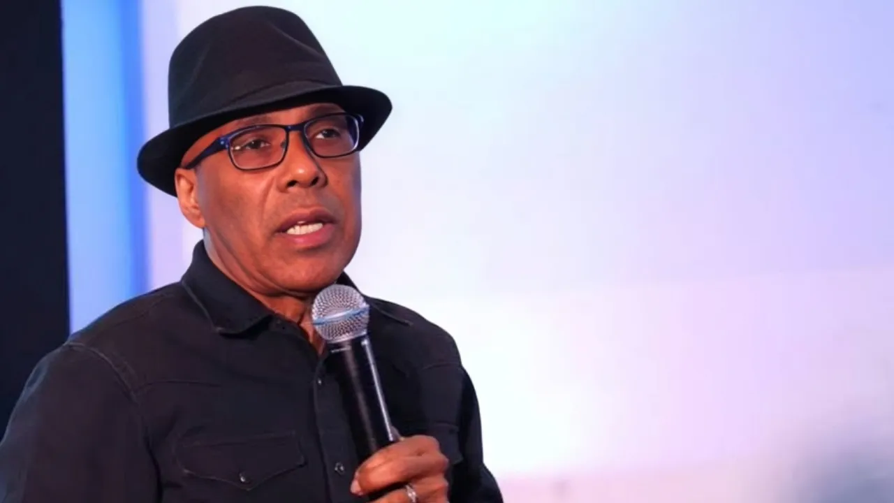 Man wearing a black fedora and glasses speaks into a handheld microphone on stage in a black shirt.