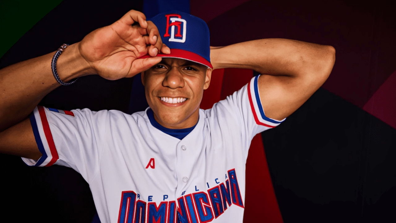 Smiling young man in a white Dominican baseball jersey and blue-red cap saluting with his hand on the brim.