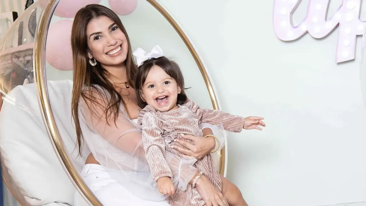 Mother sits in a round hanging chair, smiling while holding her happy young daughter in a pastel-themed room with pink decor and balloons.