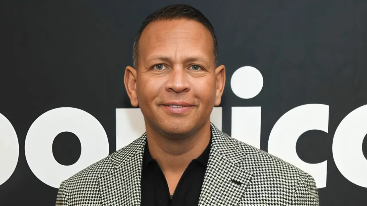 Smiling man in a black shirt and houndstooth blazer poses in front of a dark backdrop with large white text behind him.