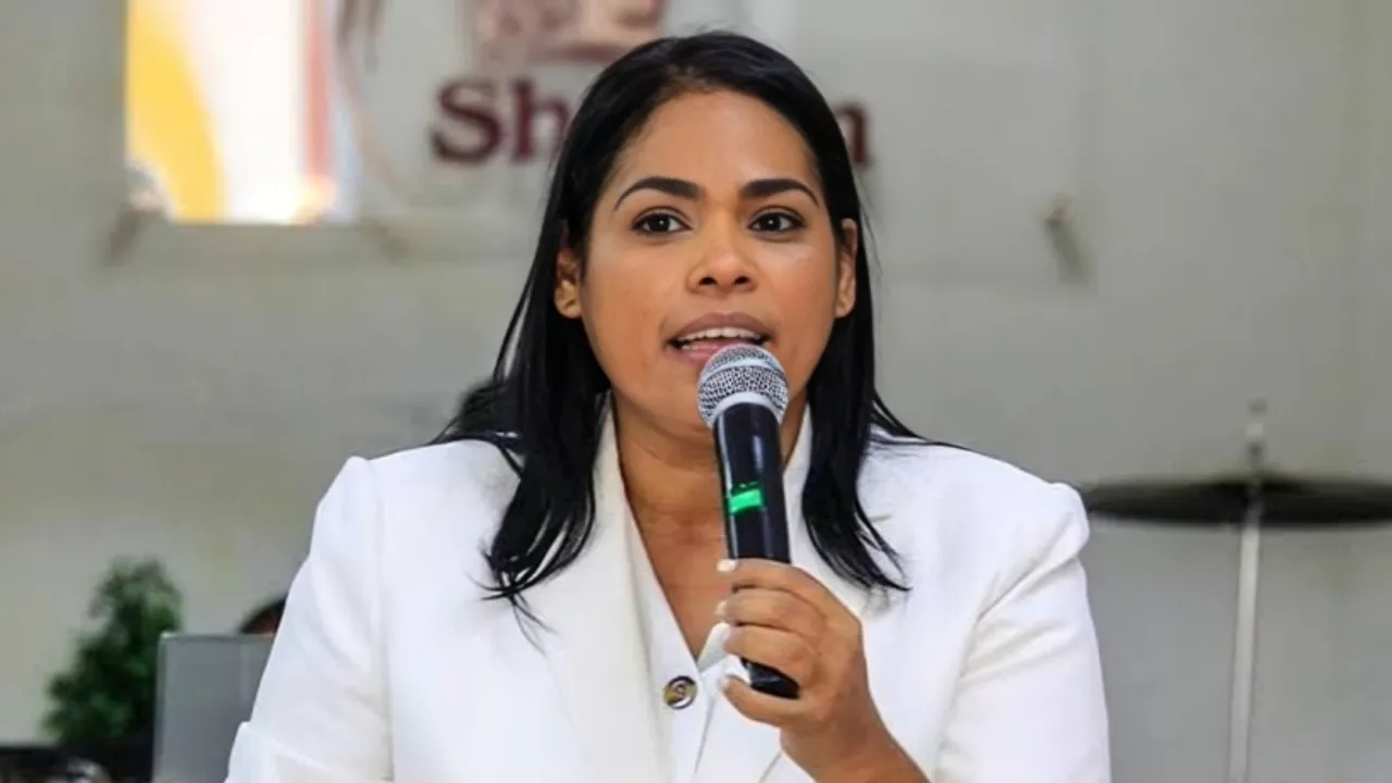 Woman in a white blazer speaks into a handheld microphone at a public event.