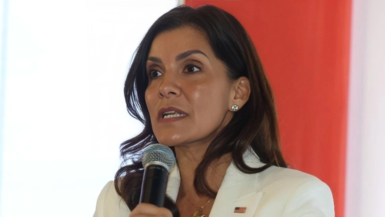 Woman in a white blazer speaks into a handheld microphone at a public event with a red backdrop.