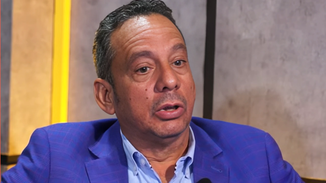 Man in a blue blazer and light blue shirt speaking in a studio setting during an interview or discussion.