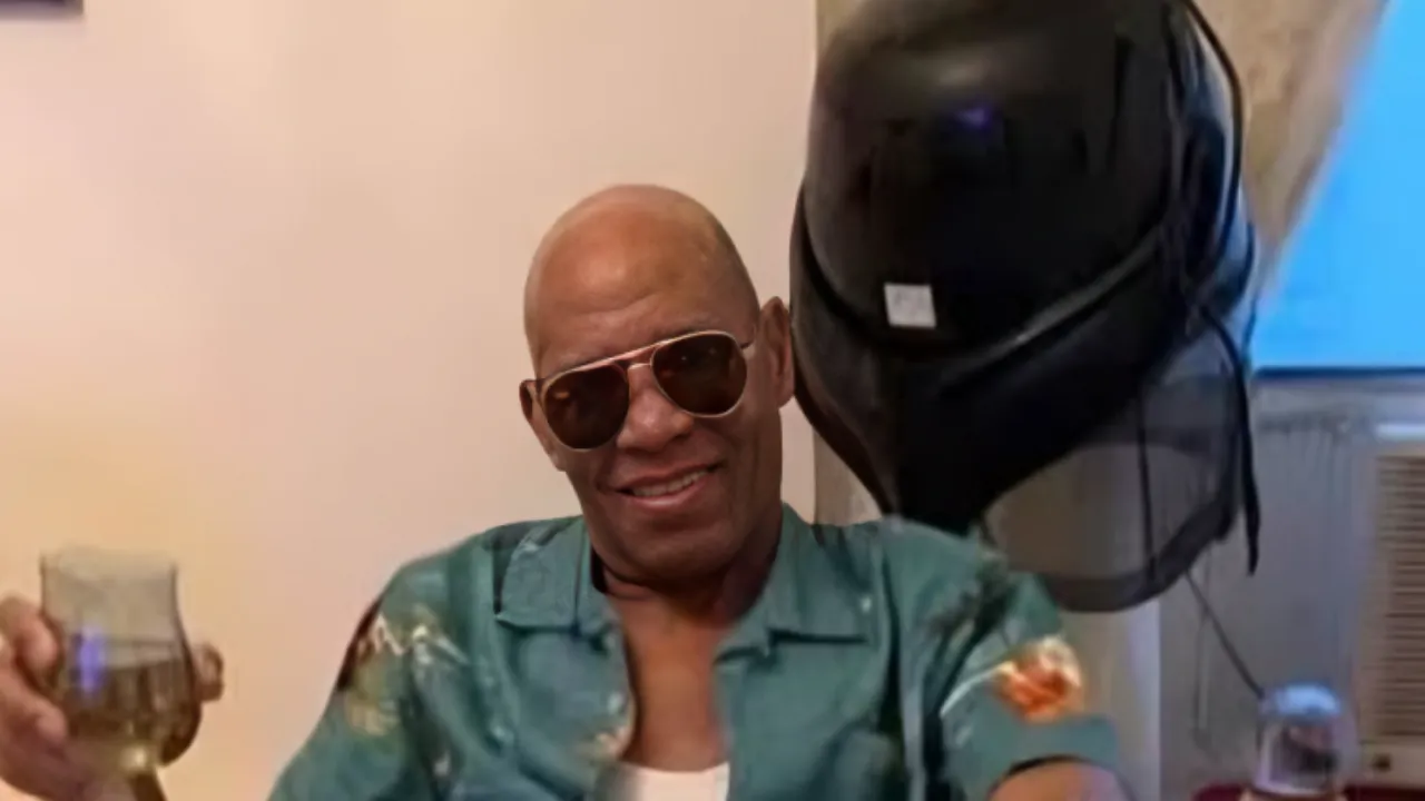 Man wearing sunglasses and a teal floral shirt raises a glass in a toast with a smile.