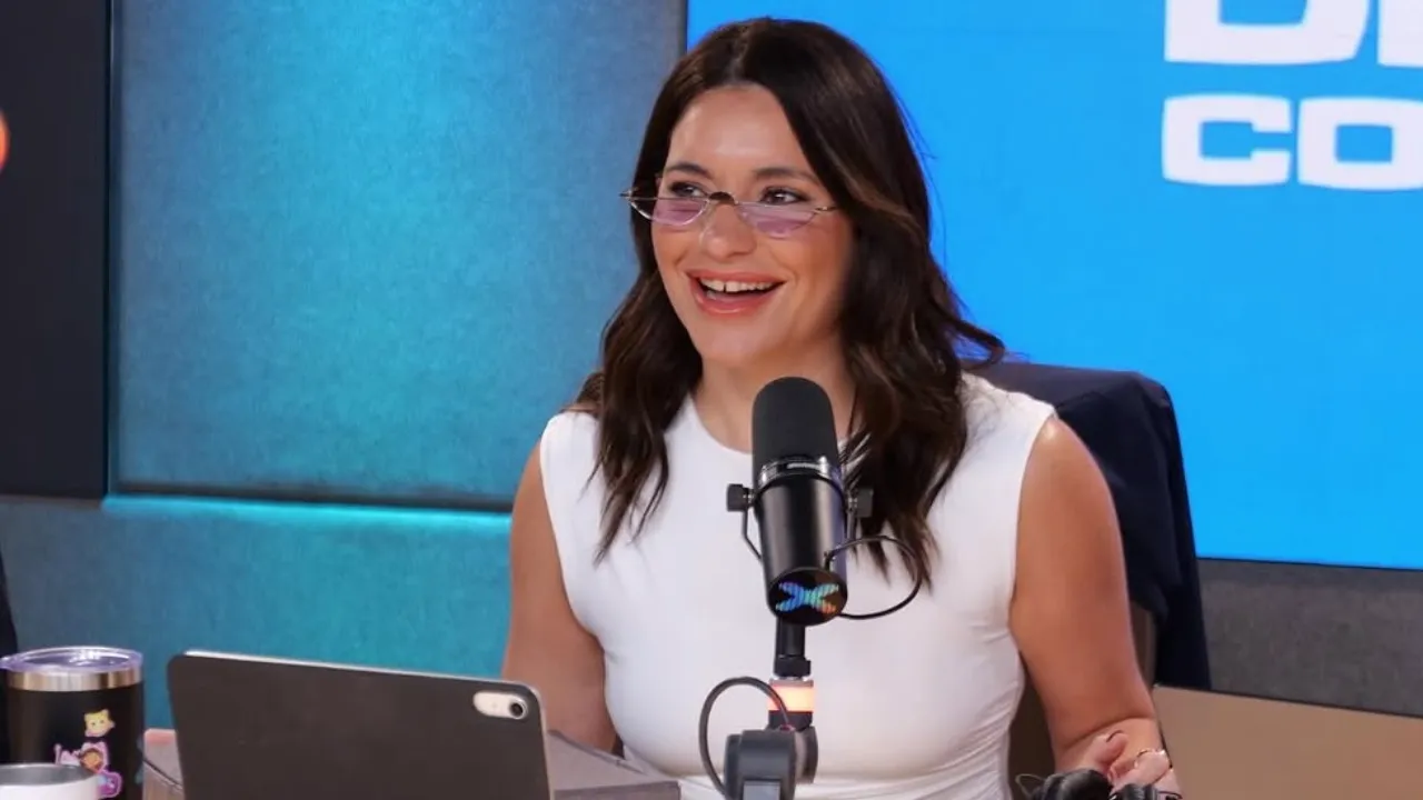 Smiling woman with dark hair and pink-tinted glasses speaks into a studio microphone with a blue backdrop.