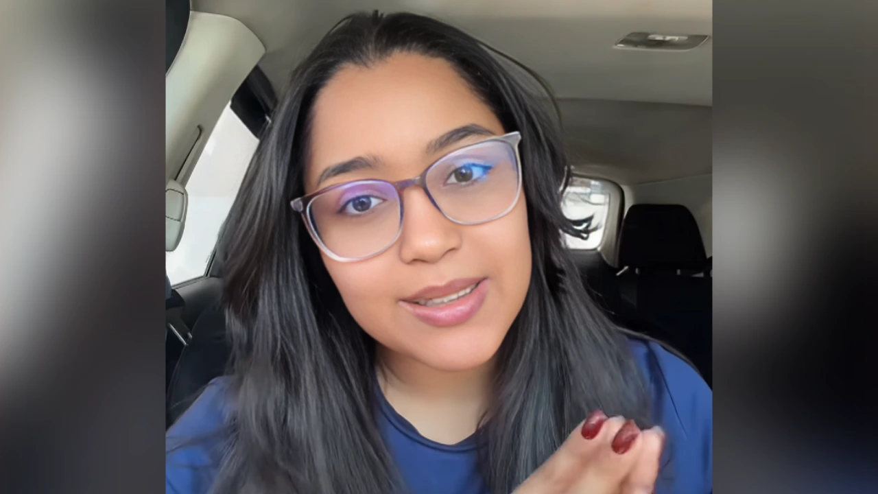 Woman with glasses speaking to camera inside a car, hands clasped, wearing a blue top, neutral expression