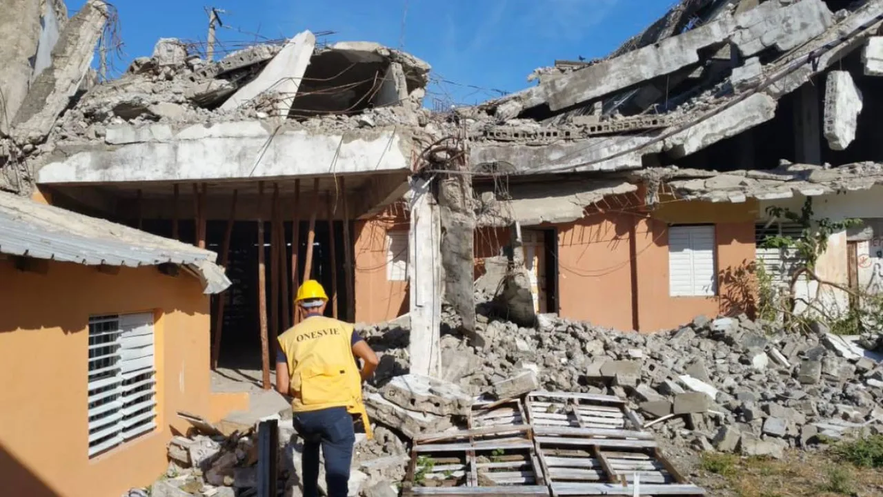 Construction worker in a yellow helmet and vest walks through rubble of collapsed buildings.