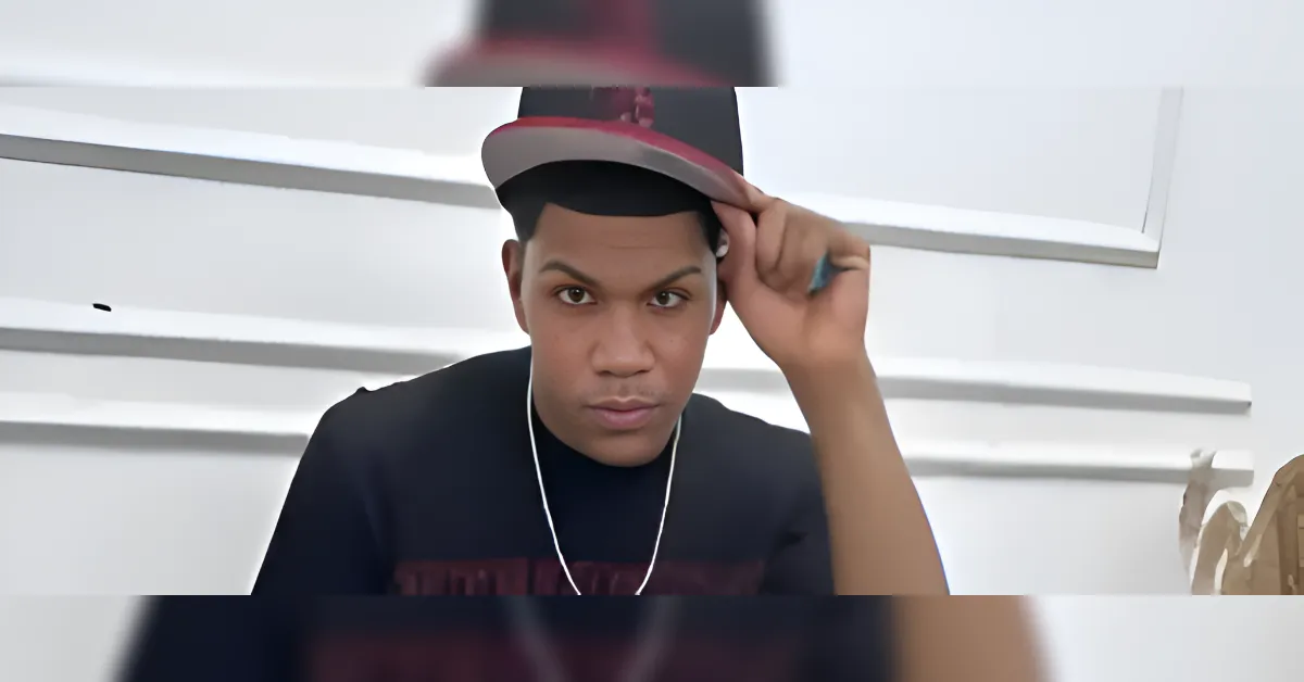 Male portrait: man in a black shirt wearing a red-and-black cap, adjusting it toward the camera, white wall behind him.