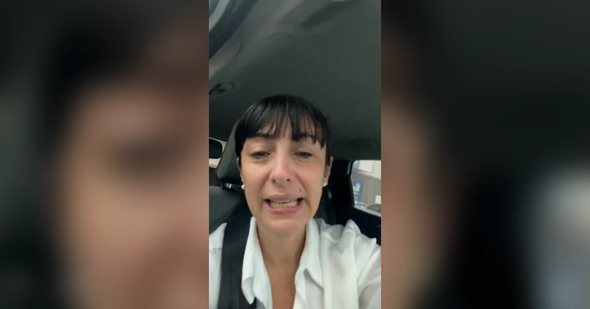 Woman wearing a white blouse speaks to the camera from inside a car, with a seatbelt across her chest.
