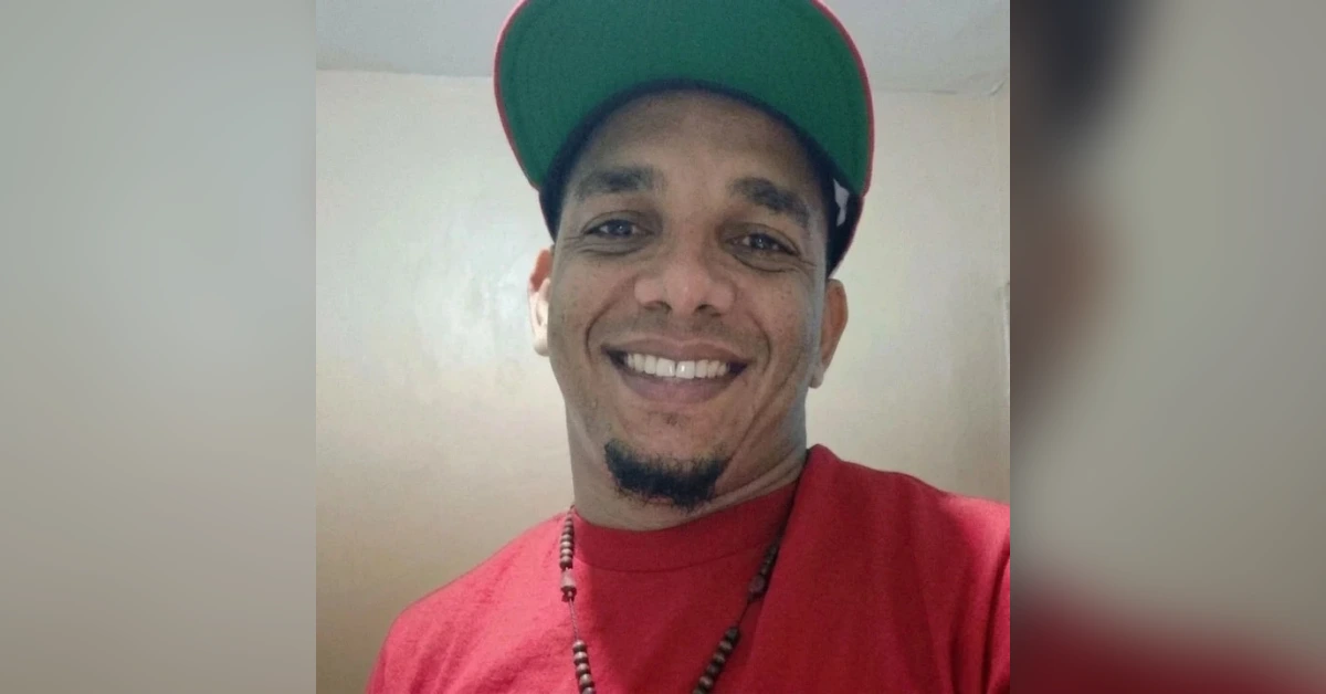 Smiling man wearing a red shirt and green cap, posing for a selfie indoors with a bead necklace visible.