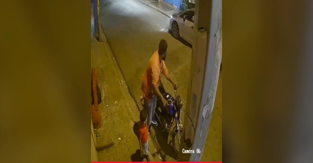 Nighttime street scene: a man in an orange shirt stands beside a blue motorcycle, with a small child nearby; ‘Camera 06’ watermark.