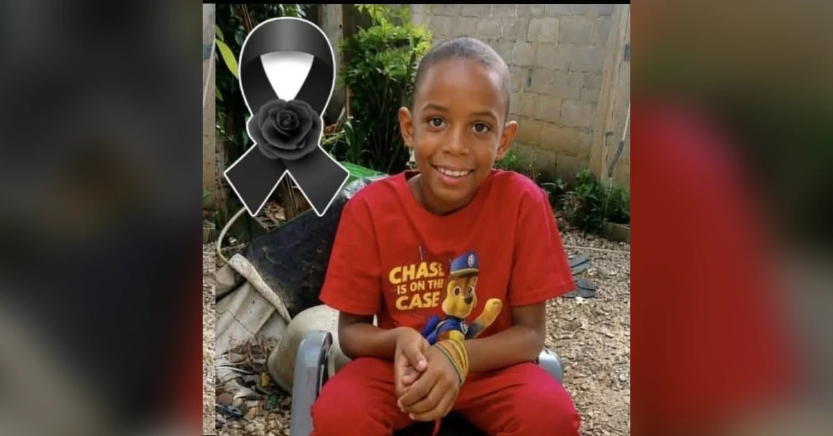 Smiling boy in a red Paw Patrol shirt sits outdoors on a plastic chair, with a black memorial ribbon graphic in the upper left.