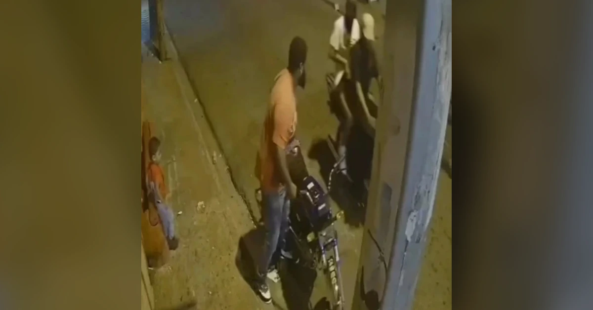 Nighttime street scene with a man in an orange shirt standing beside a parked motorcycle, while bystanders observe from the doorway nearby.
