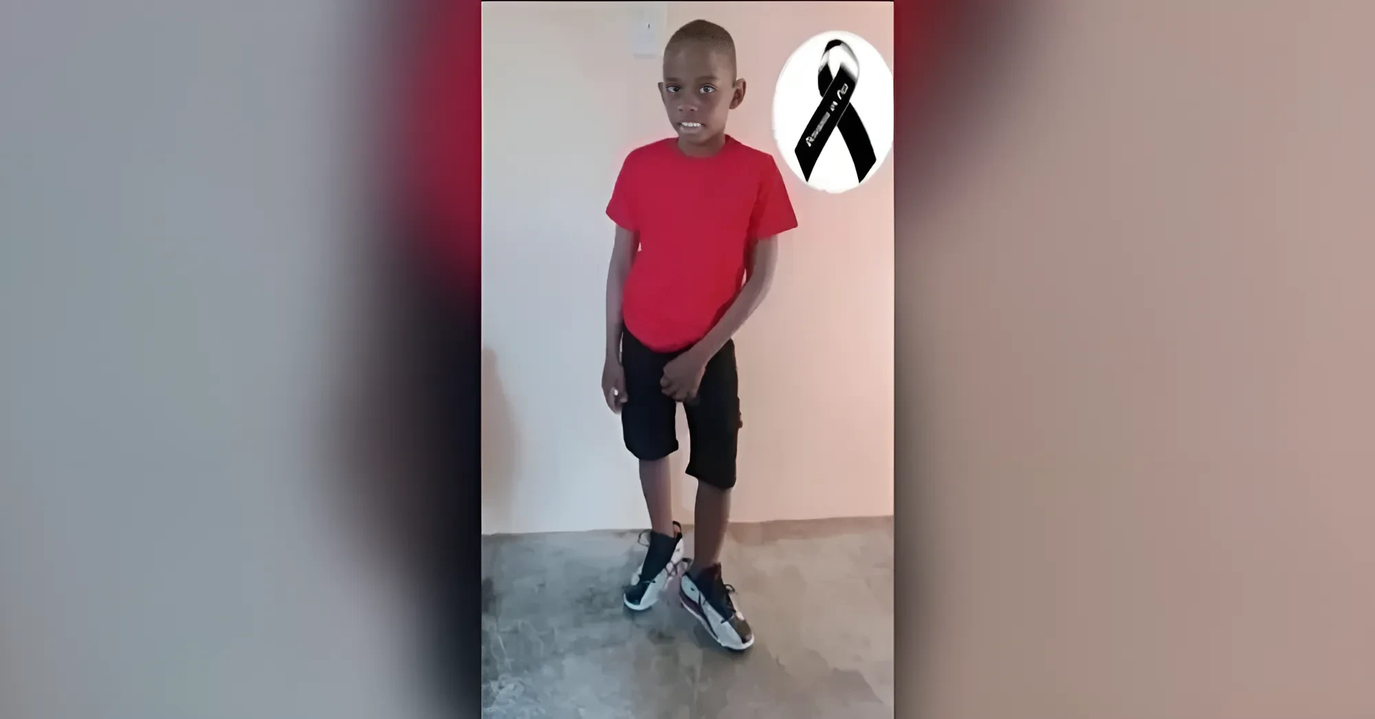 Boy in a red t‑shirt and black shorts standing indoors, sneakers on, with a black awareness ribbon graphic to his side.