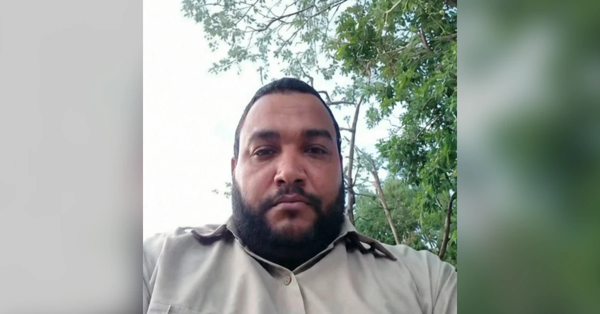Close-up portrait of a bearded man in a khaki shirt outdoors, with green trees in the background.
