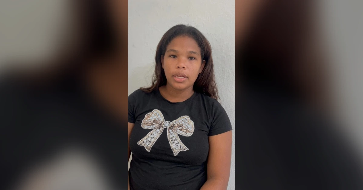 Young girl with shoulder-length dark hair wearing a black t-shirt featuring a large rhinestone bow, standing against a light gray wall.