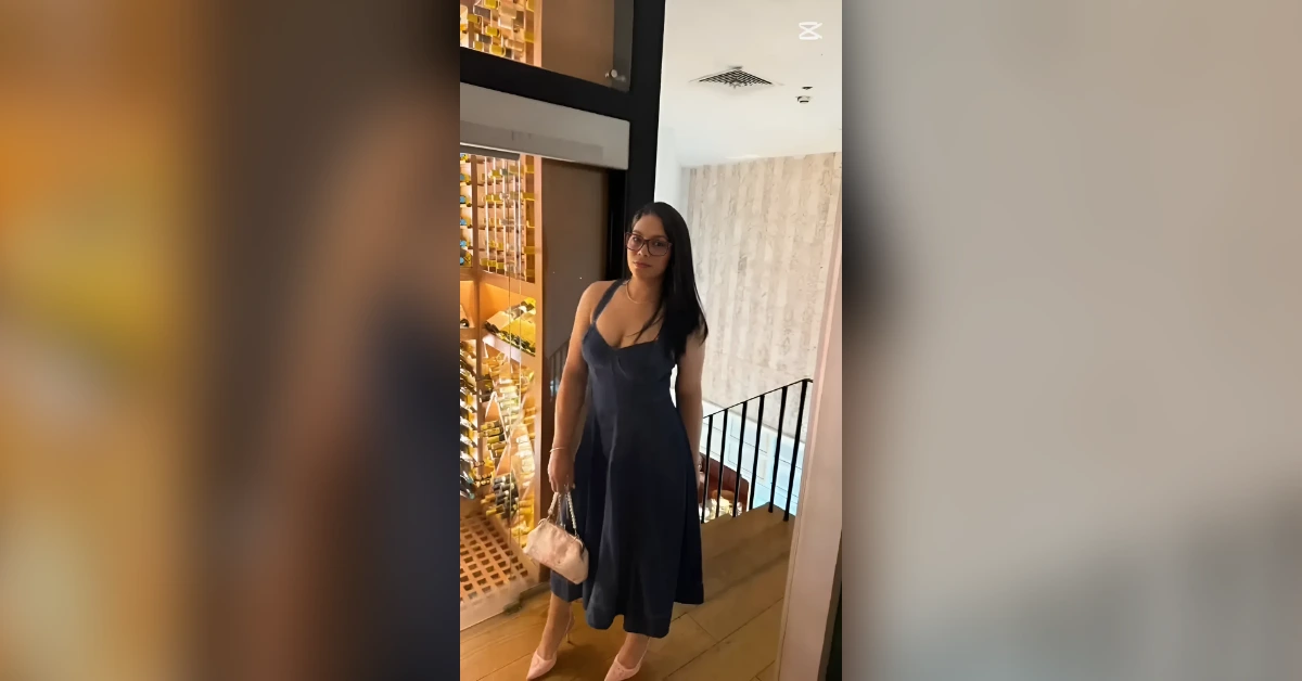 Woman in a navy spaghetti-strap midi dress stands by a wine rack in a modern interior, holding a small pink purse.