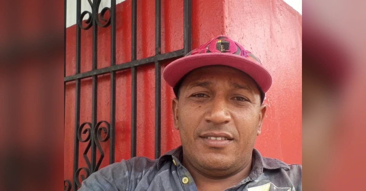 Self-portrait of a man wearing a pink cap and gray shirt, standing in front of a red wall with a black decorative gate