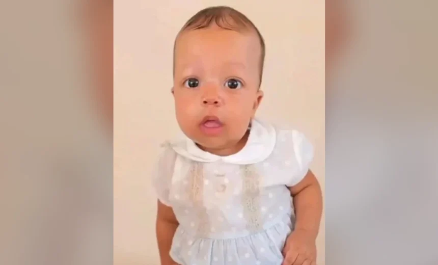 Baby wearing a white dress with lace details, looking at the camera against a neutral background.