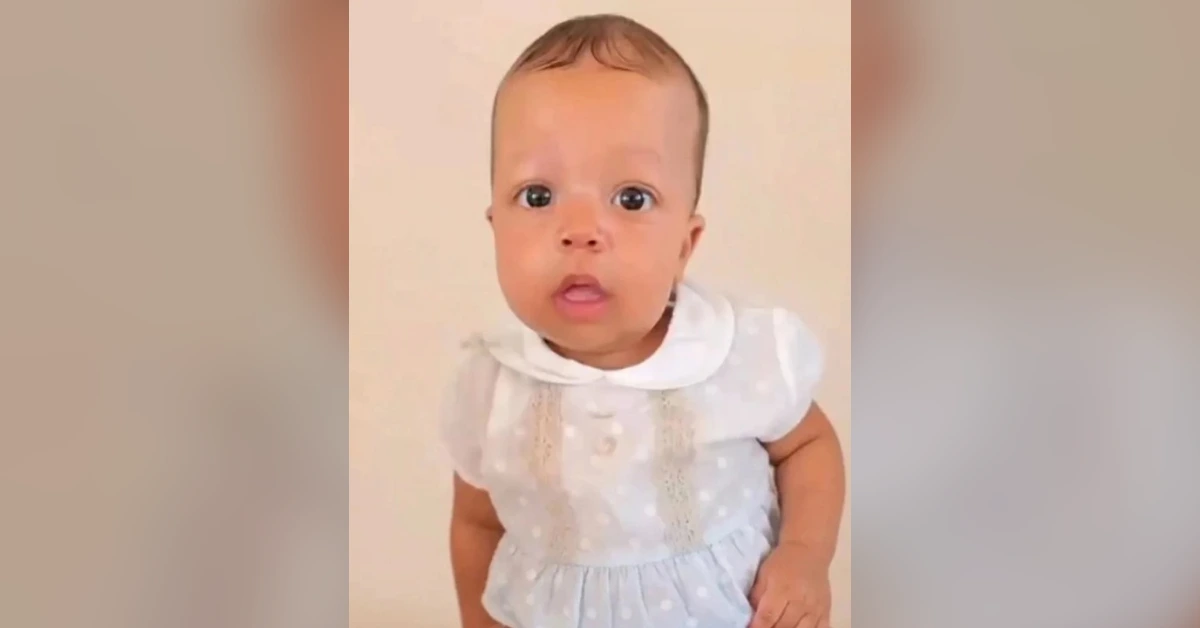 Baby girl in a light blue polka-dot dress with a white collar, looking curiously at the camera.