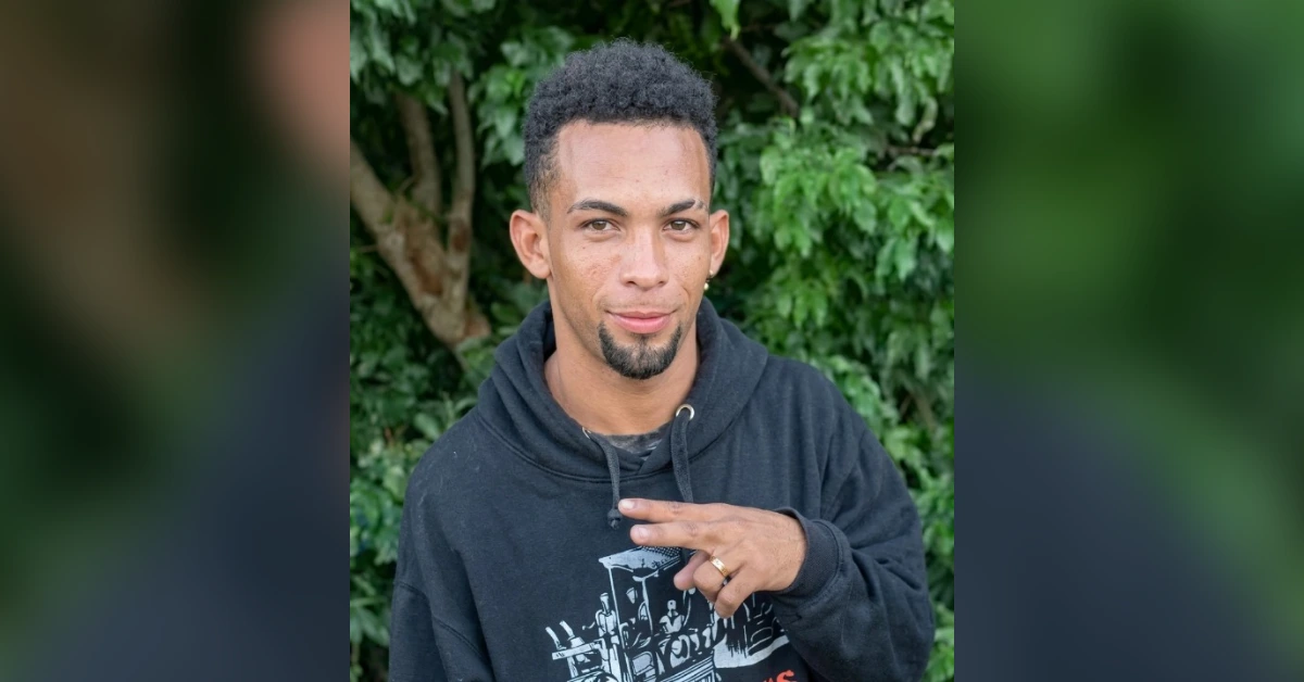 Young man in a black hoodie outdoors, making a peace sign with greenery in the background.