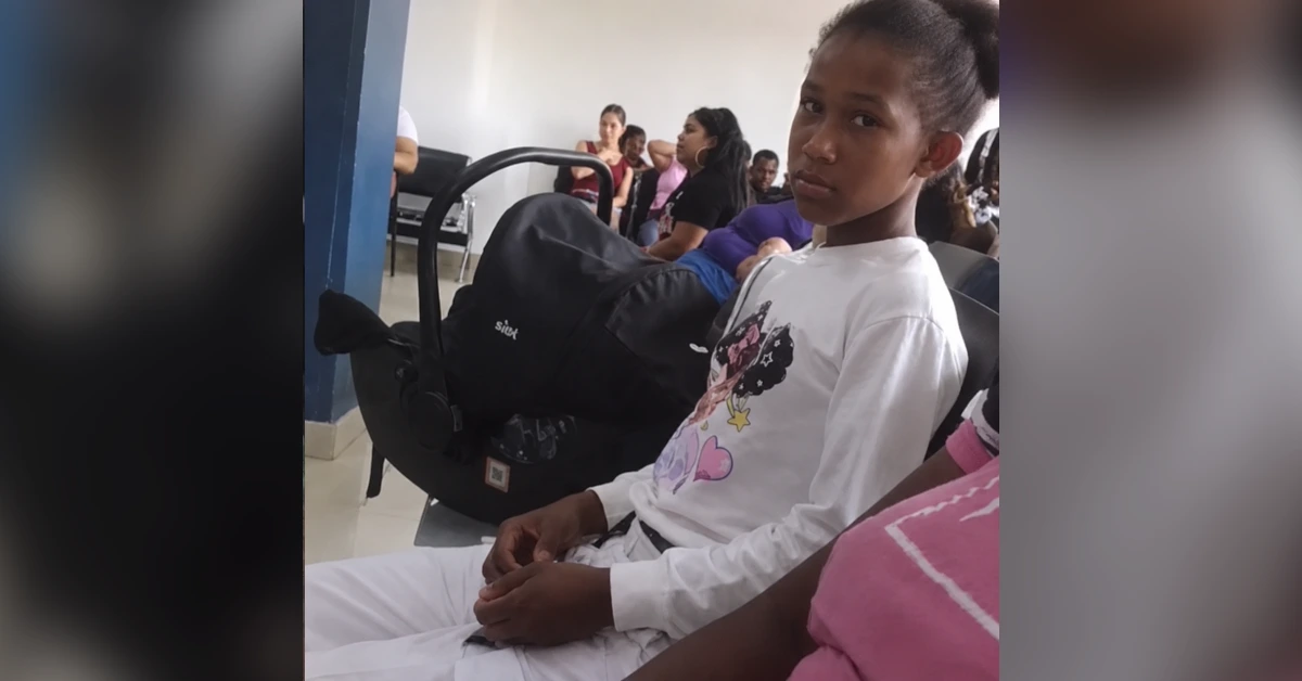 Young girl in a white shirt sits in a waiting room beside a black stroller, with people seated in the background.