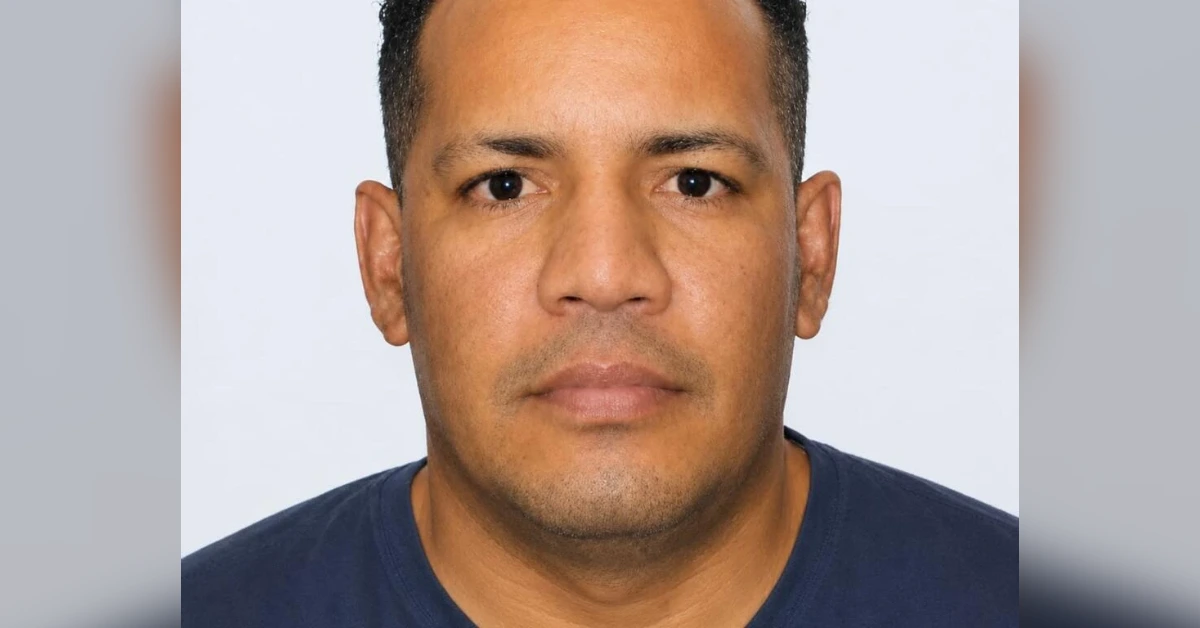 Close-up portrait of a man with short dark hair wearing a navy shirt, neutral expression, light background.
