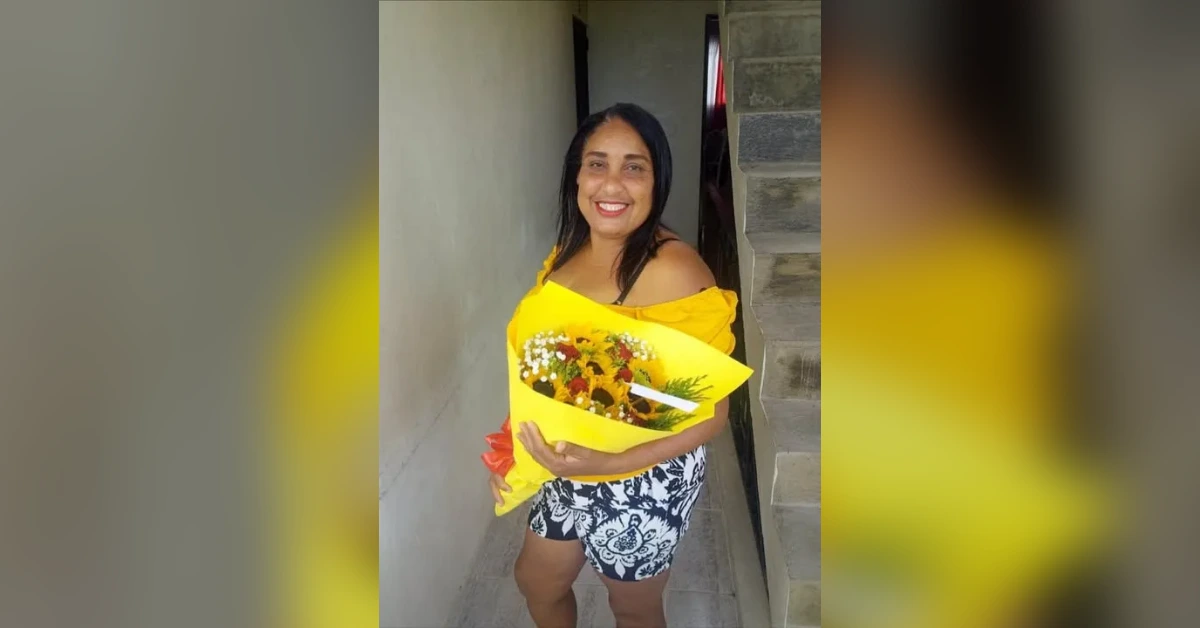 Smiling woman in a yellow off-shoulder top holding a bouquet wrapped in bright yellow paper outdoors near a concrete stairwell.