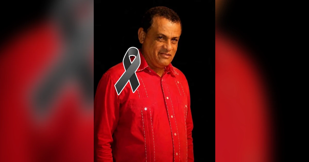 Portrait of a man in a red shirt standing against a dark background with a black memorial ribbon overlay on his chest.