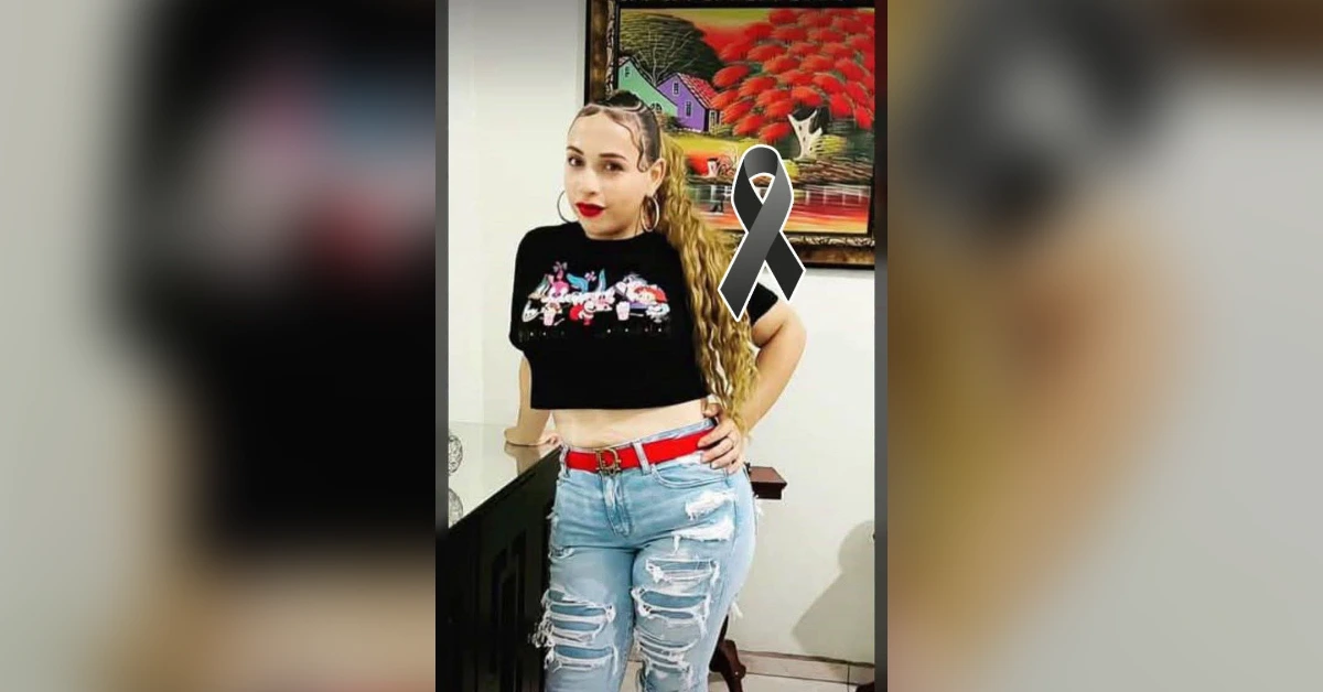 Woman posing indoors in a black cropped top and ripped jeans with a red belt; a gray awareness ribbon is overlaid on the image to indicate support or awareness.