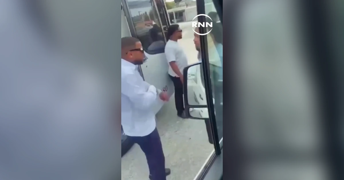 Two men stand near an open bus doorway during a confrontation: one in a white shirt and sunglasses, the other in a white T-shirt, nearby onlookers visible in the background.