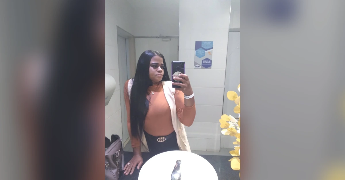Woman taking a mirror selfie in a bathroom, wearing a tan top, light vest, and black belt, with long dark hair.