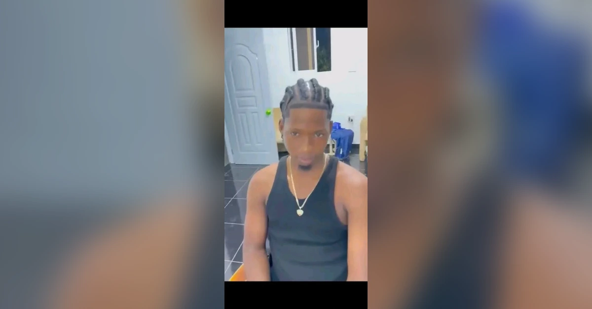 Young man with cornrow braids wearing a black sleeveless shirt and a gold necklace sits indoors, with a door and window behind him.