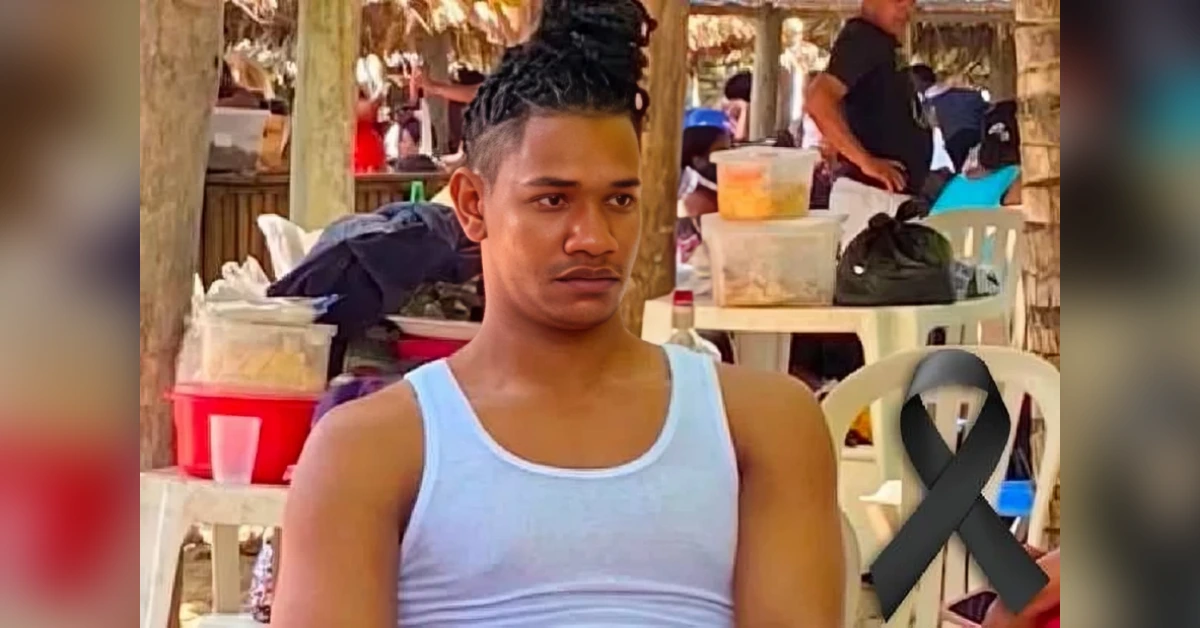 Young man in a light blue tank top sits at an outdoor beachside gathering; a black mourning ribbon is overlaid on the scene.